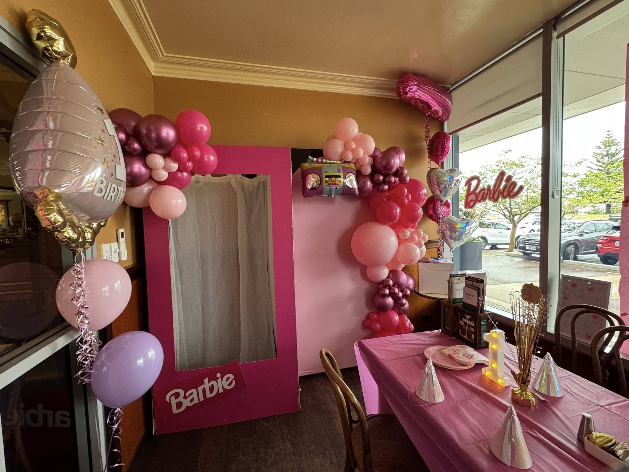A Barbie-themed birthday party setup featuring a lifesize Barbie box stand-in, pink balloons, and party decor.