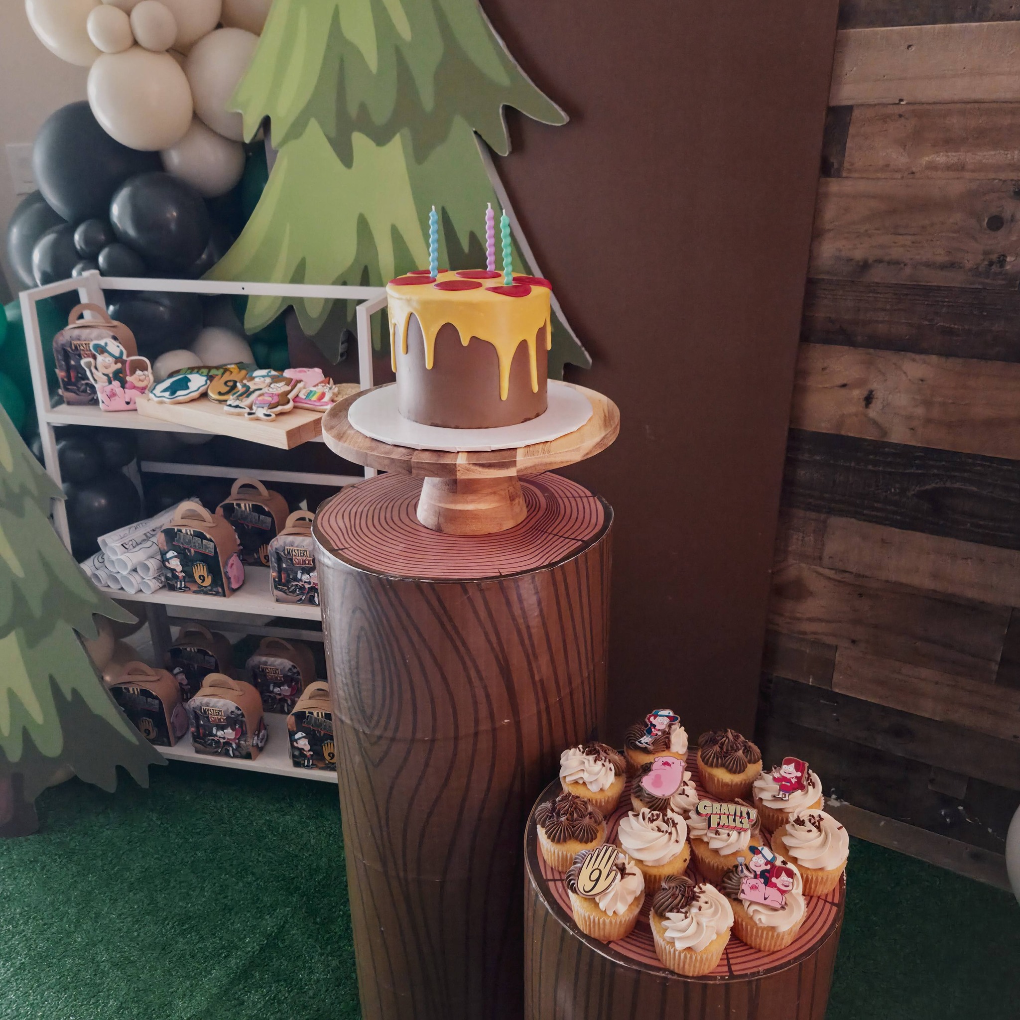 Gravity Falls themed birthday cake with yellow drip icing and matching cupcakes on wooden tree stump displays.