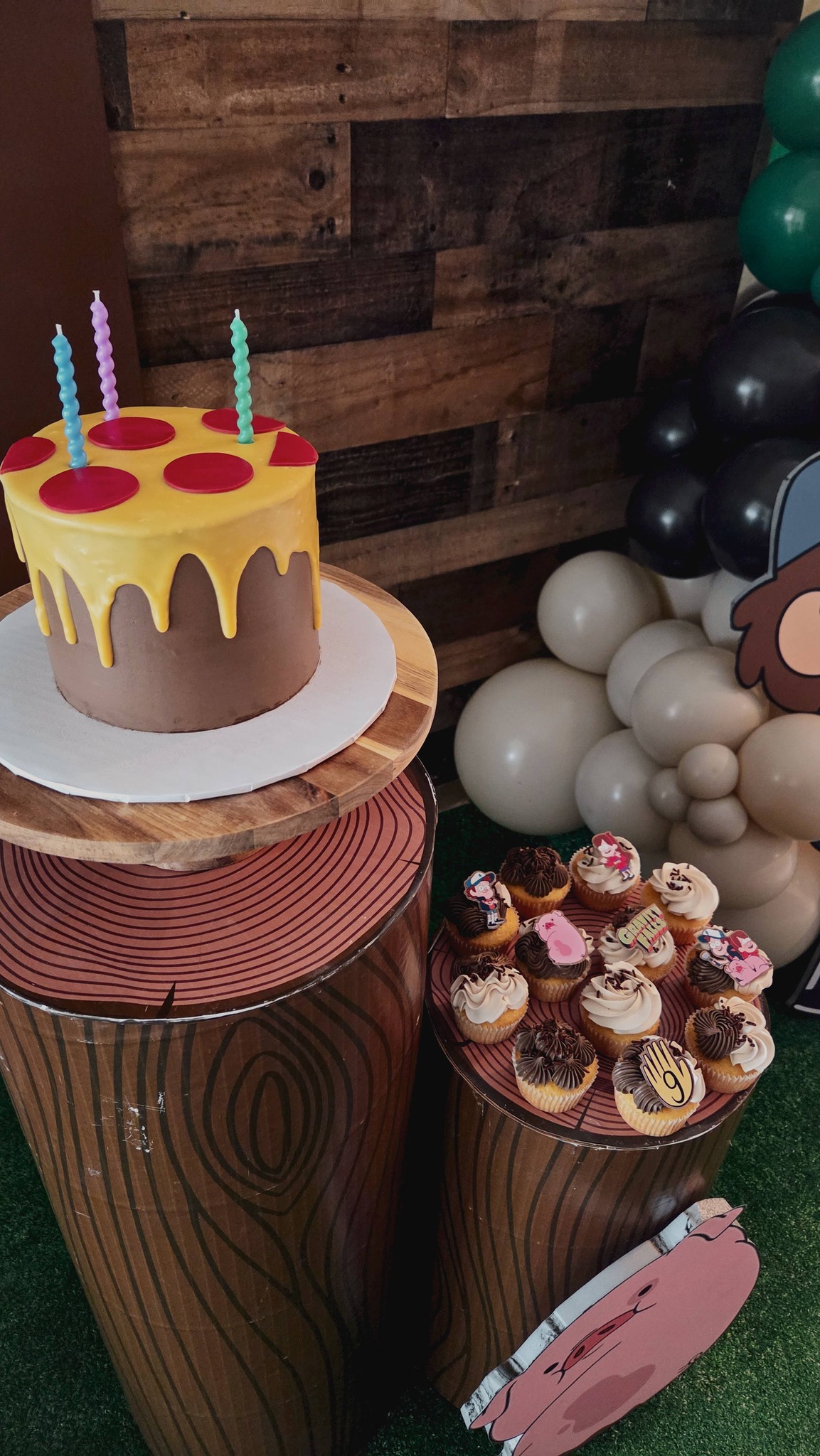 Gravity Falls themed birthday cake with yellow drip and cupcakes on tree stump pedestals.