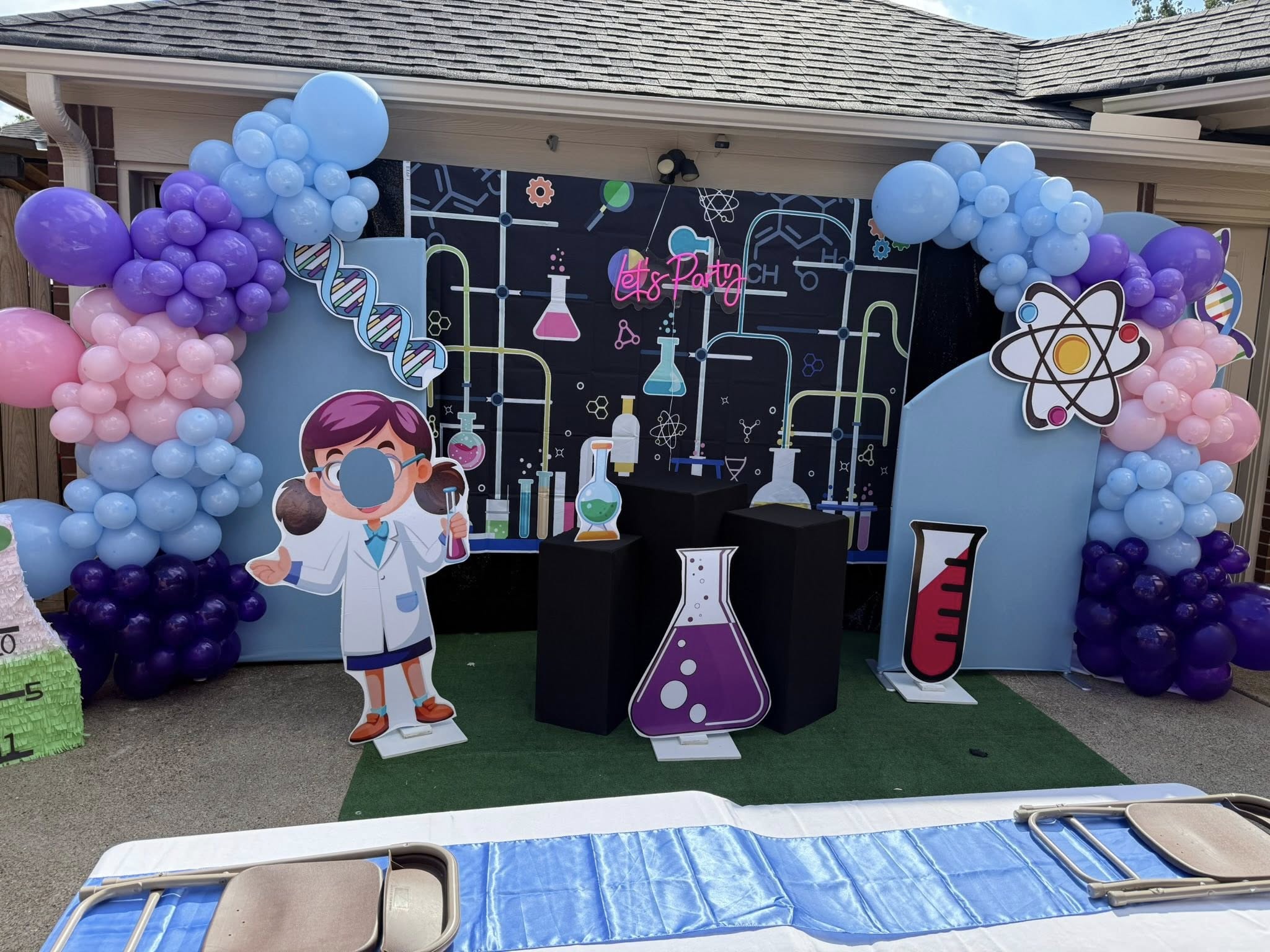 Mad scientist themed party backdrop with balloons, lab supply cutouts, and a character photo cutout.