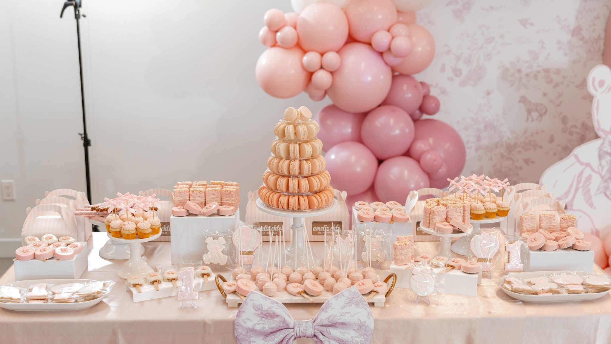 Table set with blush pink desserts, including a macaron tower, cakepops, and cookies, decorated with pink balloons