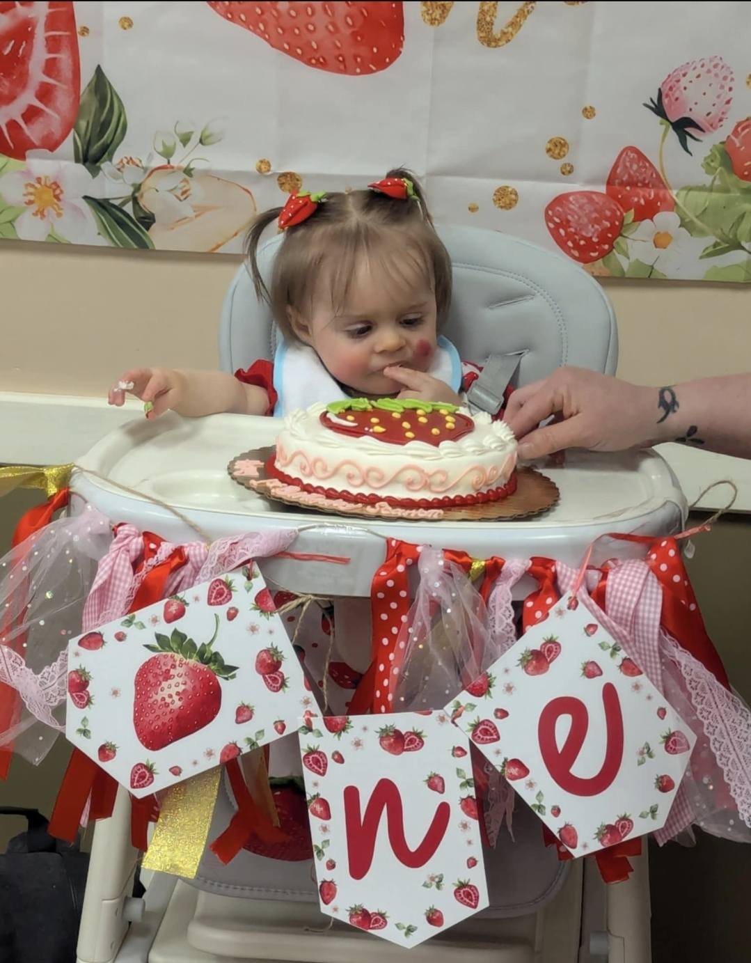 Baby celebrating first birthday in a high chair with a strawberry cake and strawberry-themed decorations.