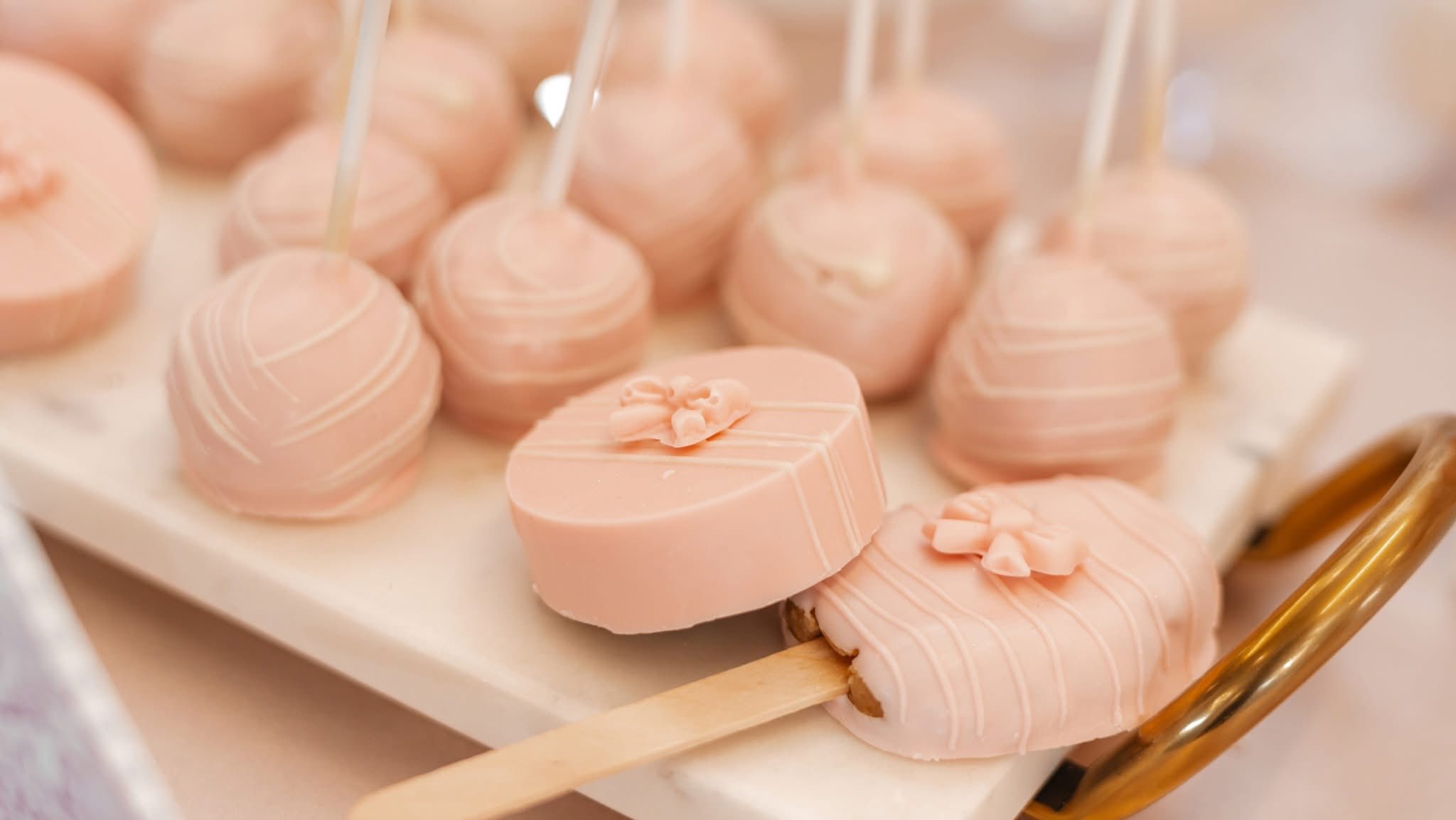 Pink cake pops and chocolate-dipped cookies decorated with bows on a marble serving tray.