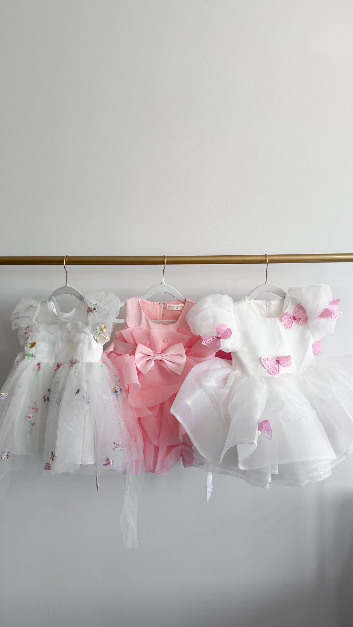 Three decorative toddler dresses with floral, ruffled, and butterfly details hanging on a gold rack