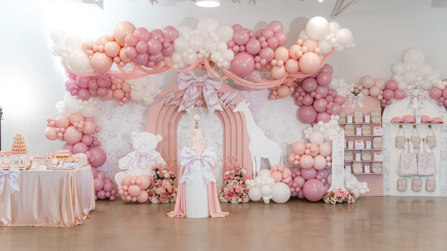 First birthday party setup featuring pink and white balloon arches, floral decor, and teddy bear and giraffe cutouts.