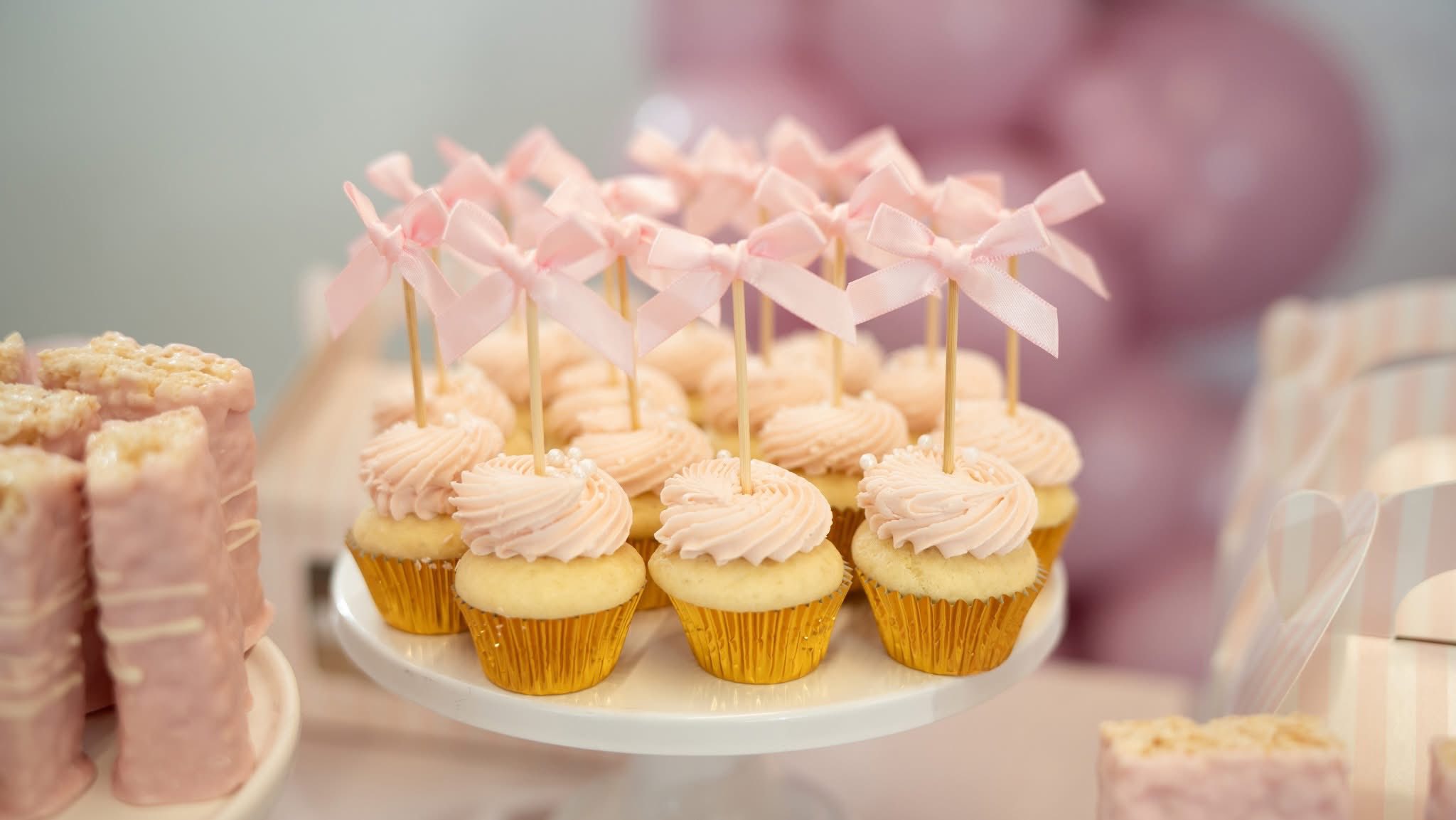 Mini cupcakes with pink frosting and decorative ribbon bow toppers on a white cake stand