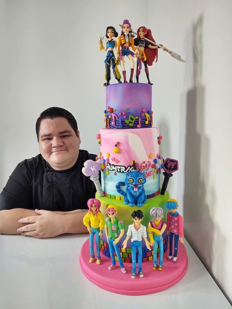 Baker posing with a multi-tiered themed cake featuring anime figures and music-inspired decorations.