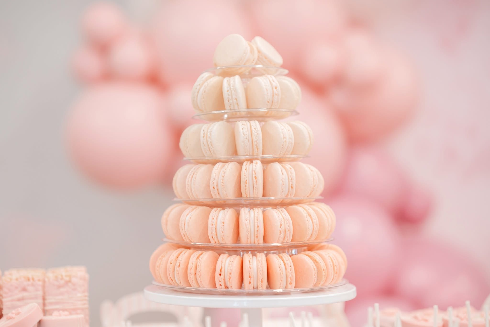 Tiered stand stacked with pale pink macarons, with blurred pink balloons in the background.