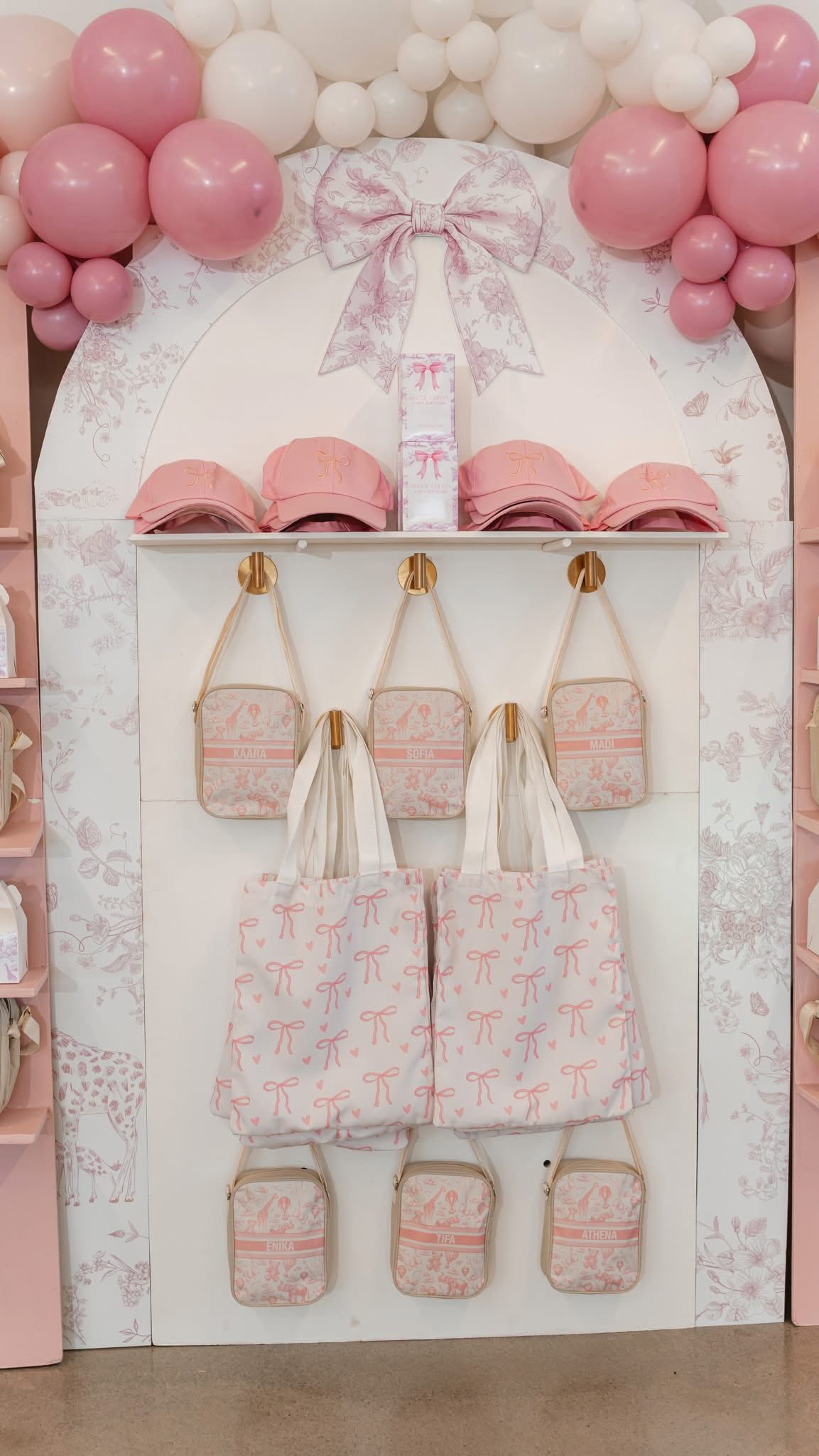 Retail display decorated with pink balloons featuring personalized bags, tote bags, and caps with bow details.