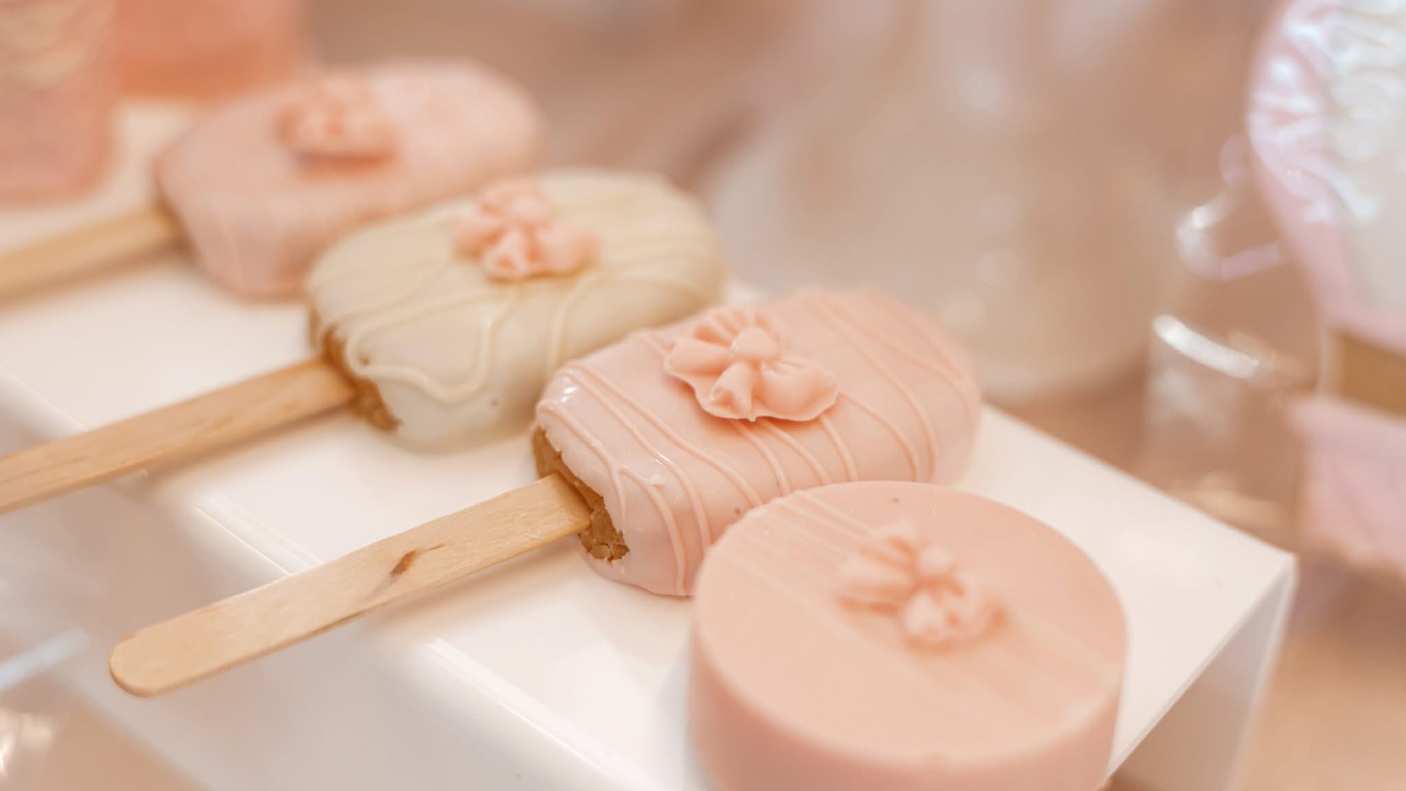 Assorted decorated cake popsicles and a cookie with soft pink and white icing and bow embellishments.