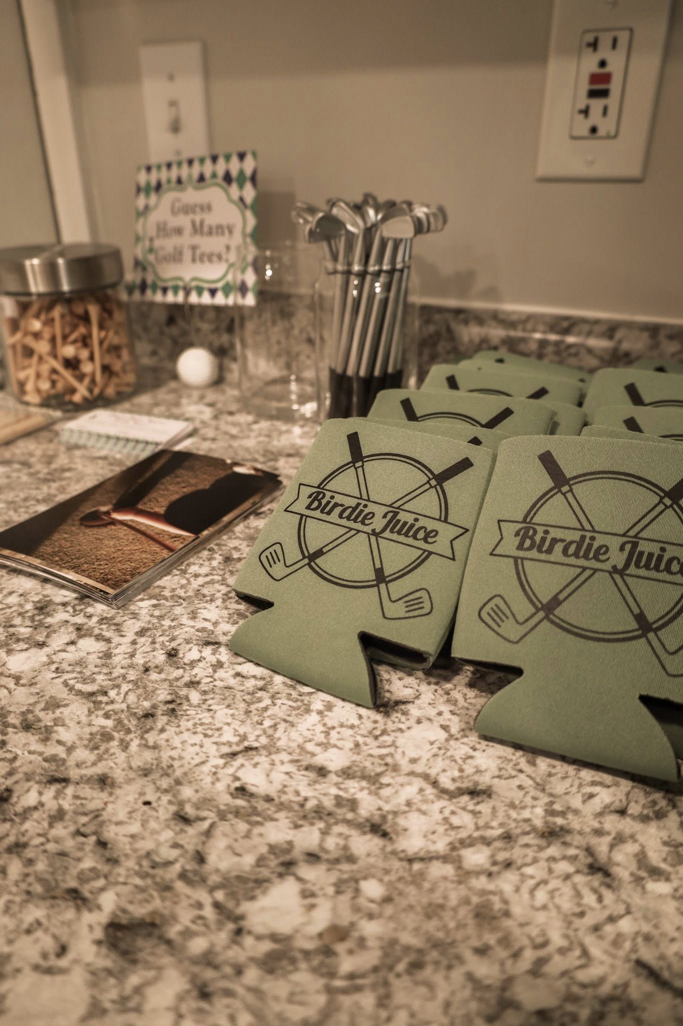 Stack of green koozies featuring a Birdie Juice golf logo with a guess the golf tees game sign in the background.