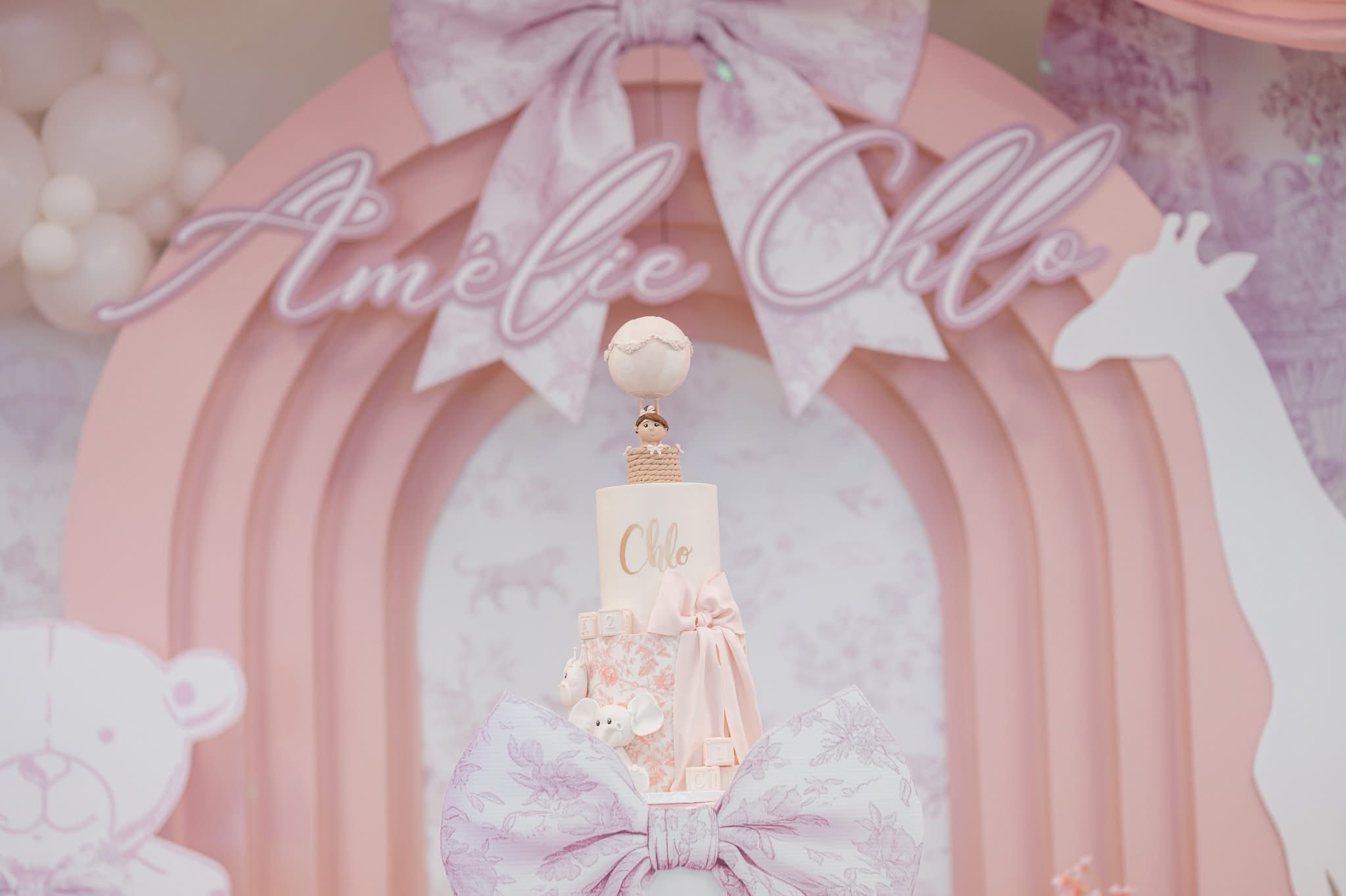 Tiered birthday cake with hot air balloon topper and elephant accents in front of a pink Amélie Chlo display.