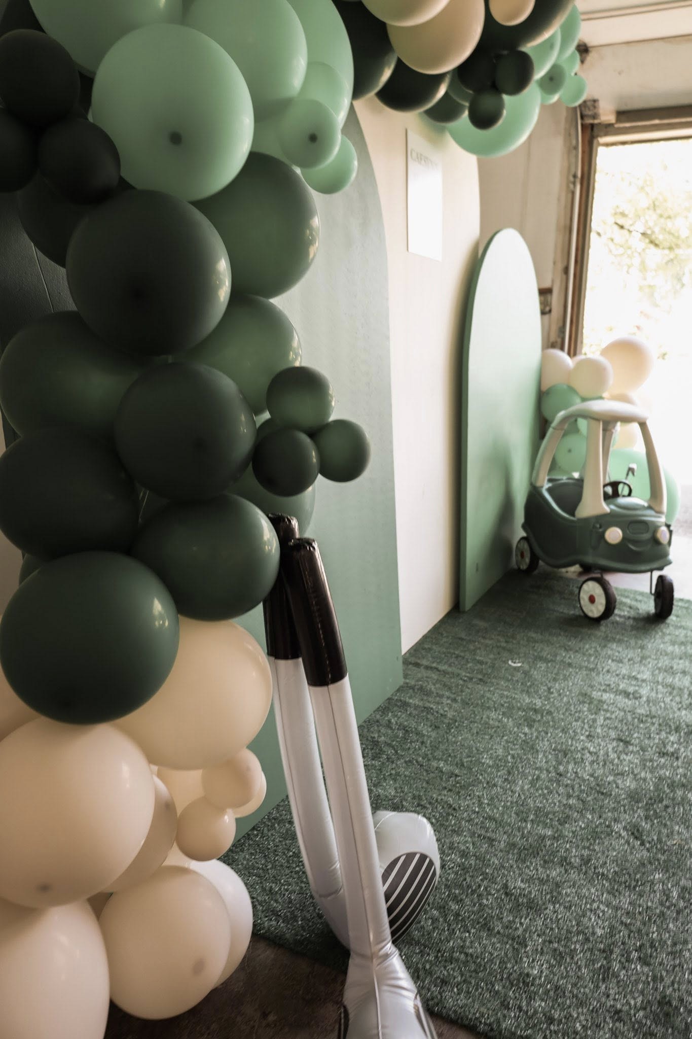 Sage green and cream balloon arch with a toy car decoration for a children's party setup.