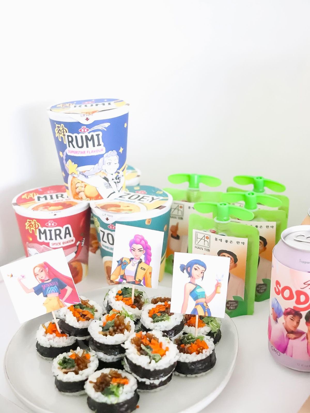 A plate of kimbap rolls topped with decorative character paper picks, surrounded by Rumi noodles and green drink pouches.