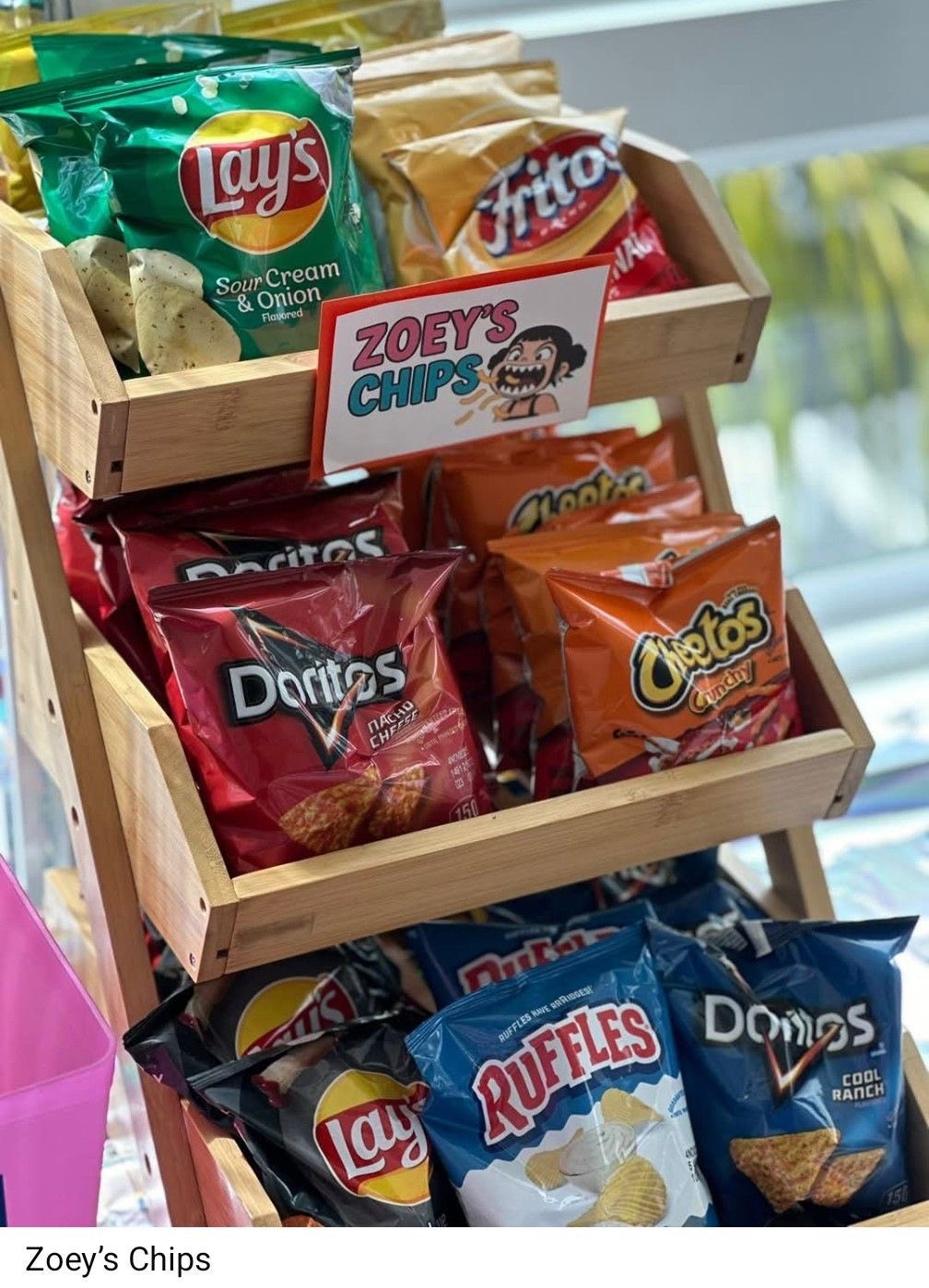 A wooden display rack filled with various snack bags including Lay's Sour Cream and Onion, Fritos, Doritos, Cheetos, and Ruffles, featuring a sign labeled Zoey's Chips.