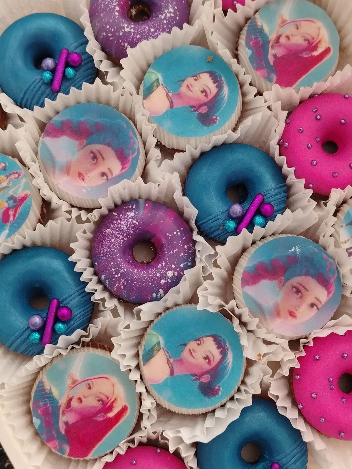 Assorted gourmet donuts featuring character images, metallic blue glaze, and glittery purple icing in a box