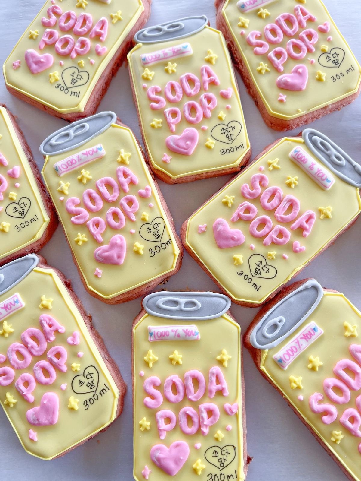 Group of soda can-shaped cookies decorated with yellow icing, pink text, and small hearts