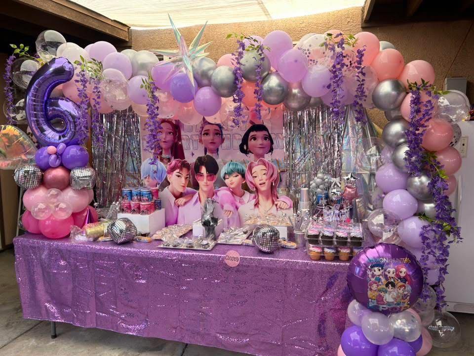Table set up for a sixth birthday party with a purple sequin cloth, K-pop style decorations, and balloon arch.