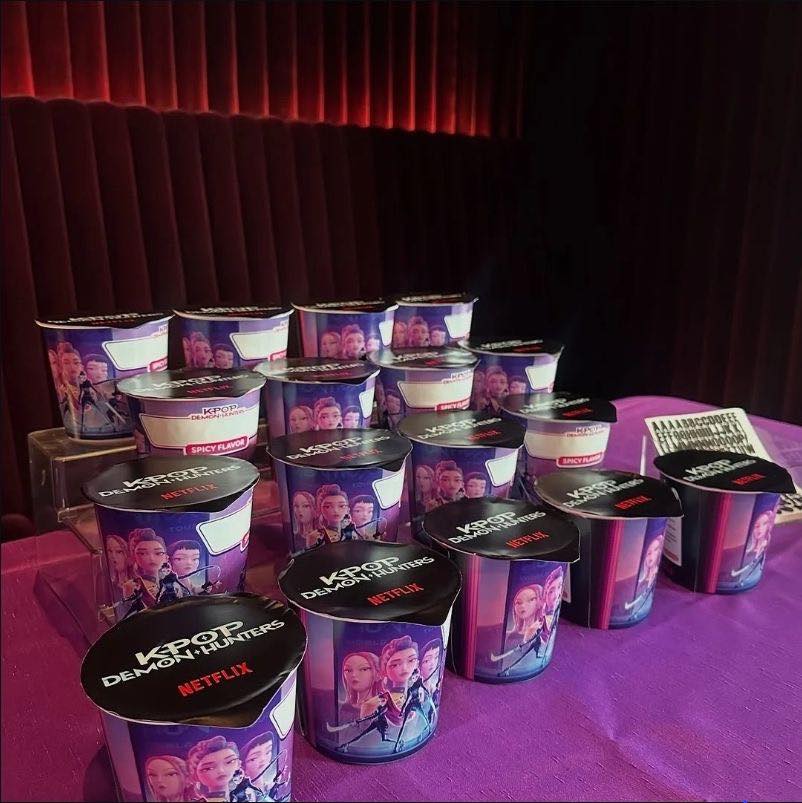 Table arranged with multiple K-Pop Demon Hunters Netflix branded spicy snack cups on a bright purple tablecloth.