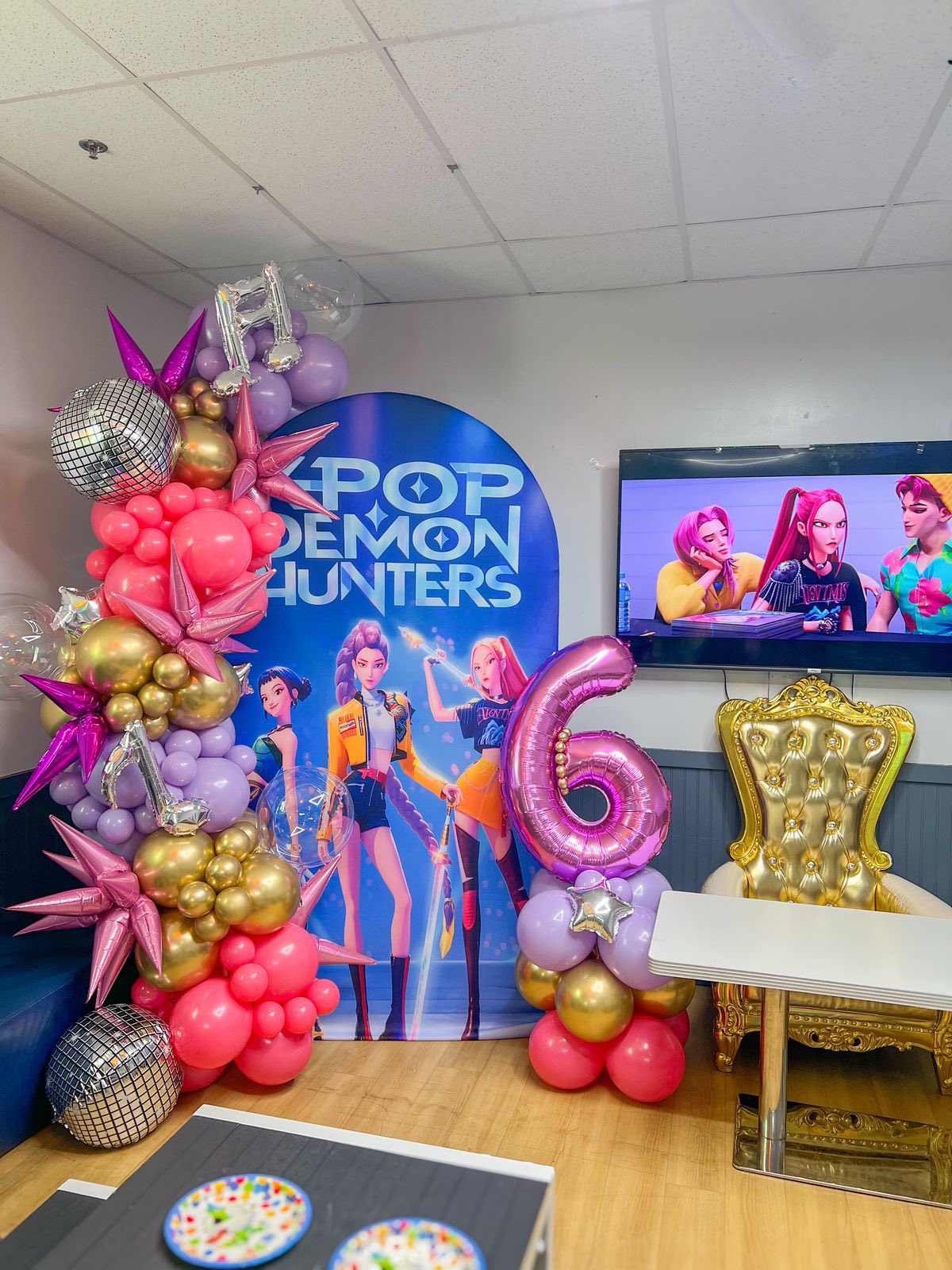 K-pop Demon Hunters themed birthday party decorations with a balloon arch, a number 6 balloon, and a gold throne.