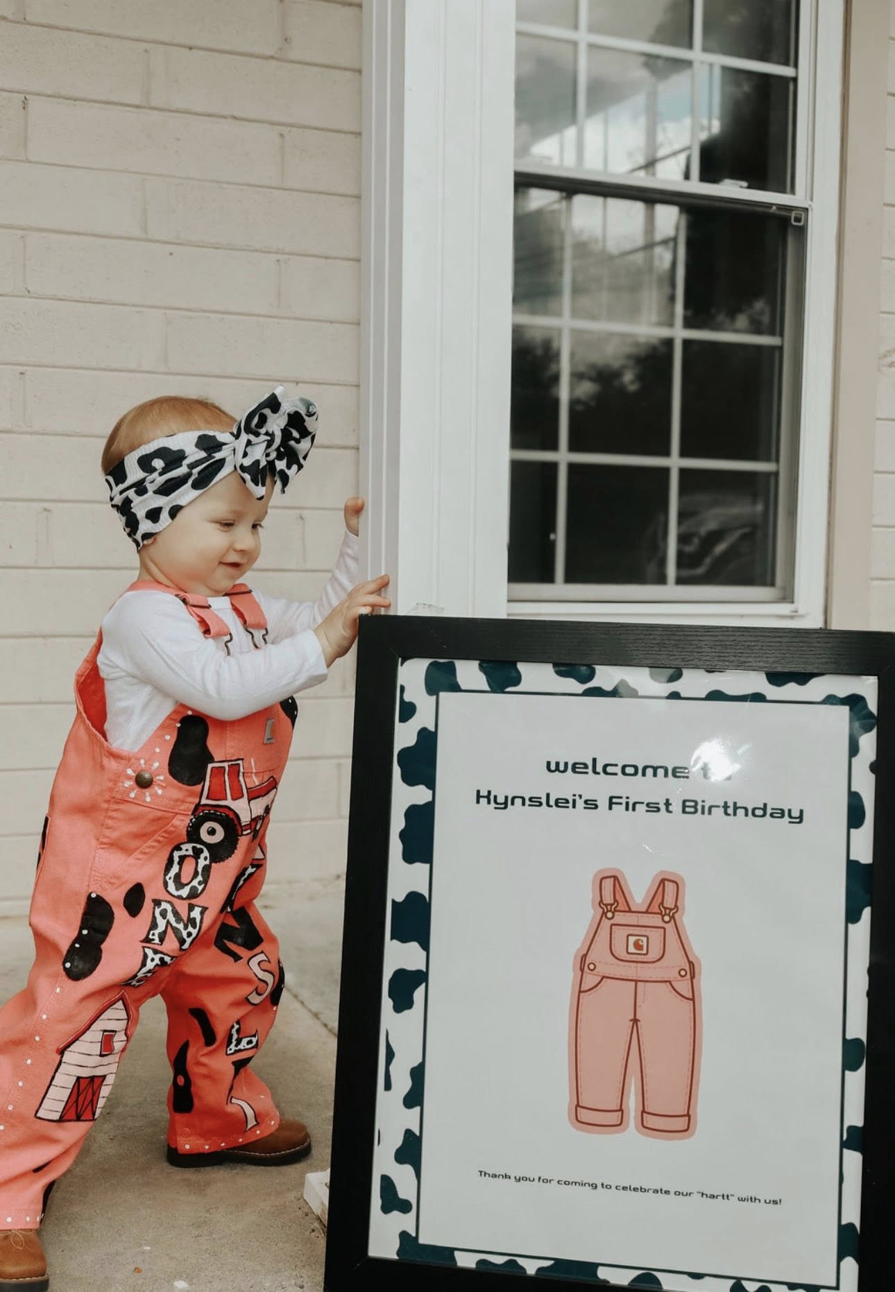 Toddler wearing pink overalls and a cow-print headband standing next to a welcome sign for a first birthday party.