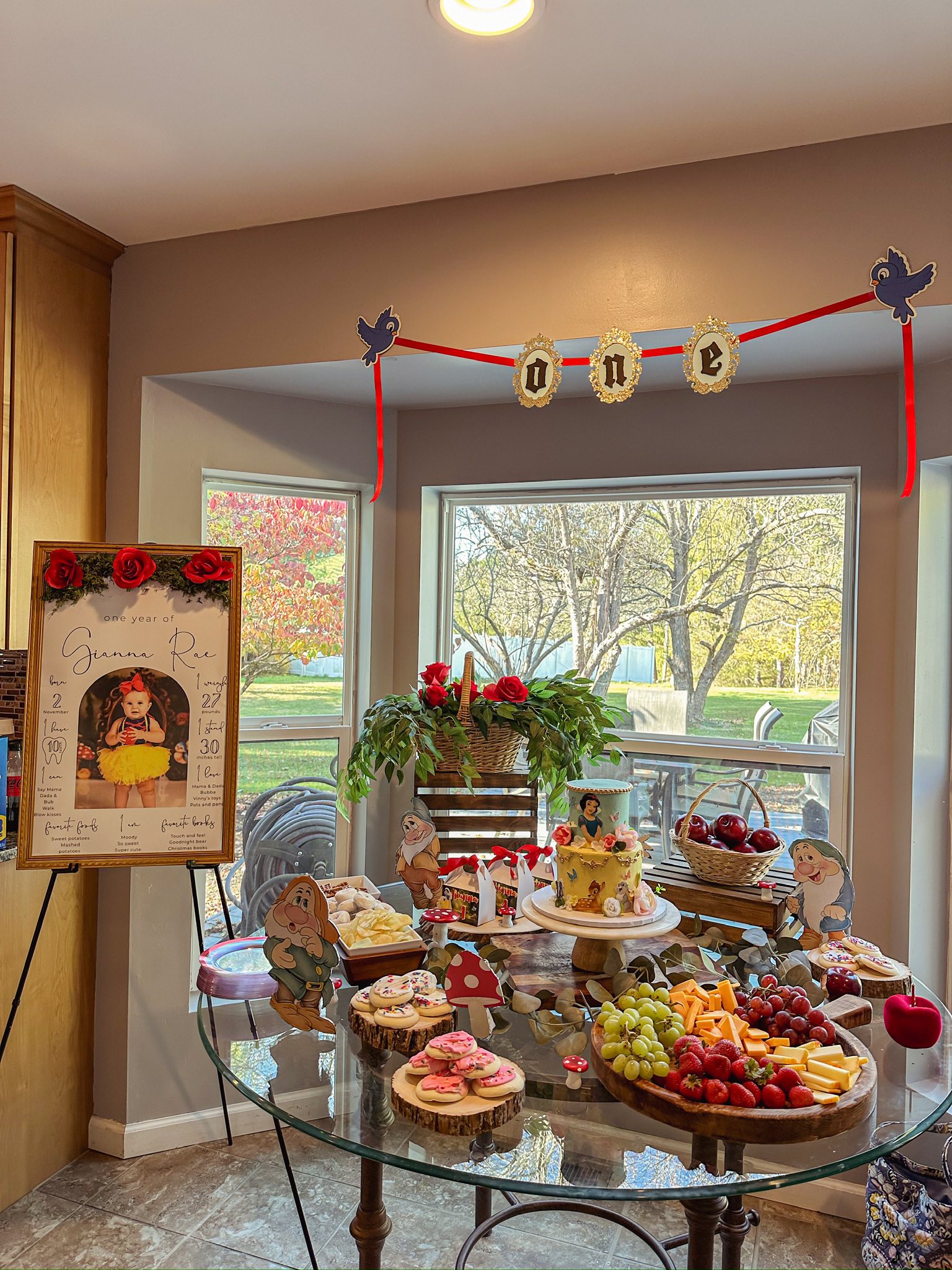Snow White themed first birthday party dessert table with a themed cake, fruit platter, and milestone sign.
