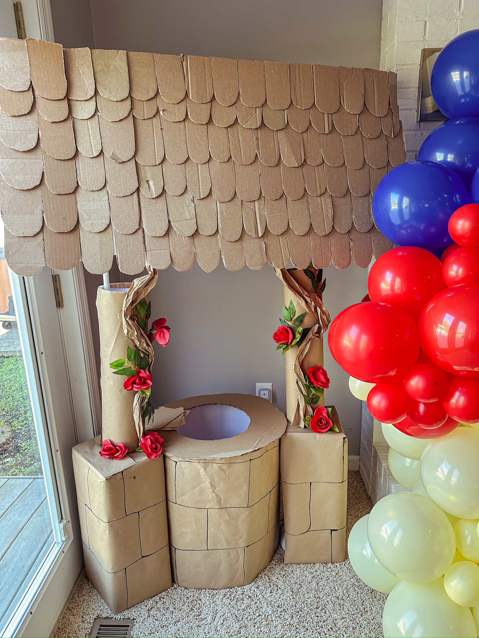 DIY cardboard wishing well prop decorated with faux roses and a colorful balloon arch