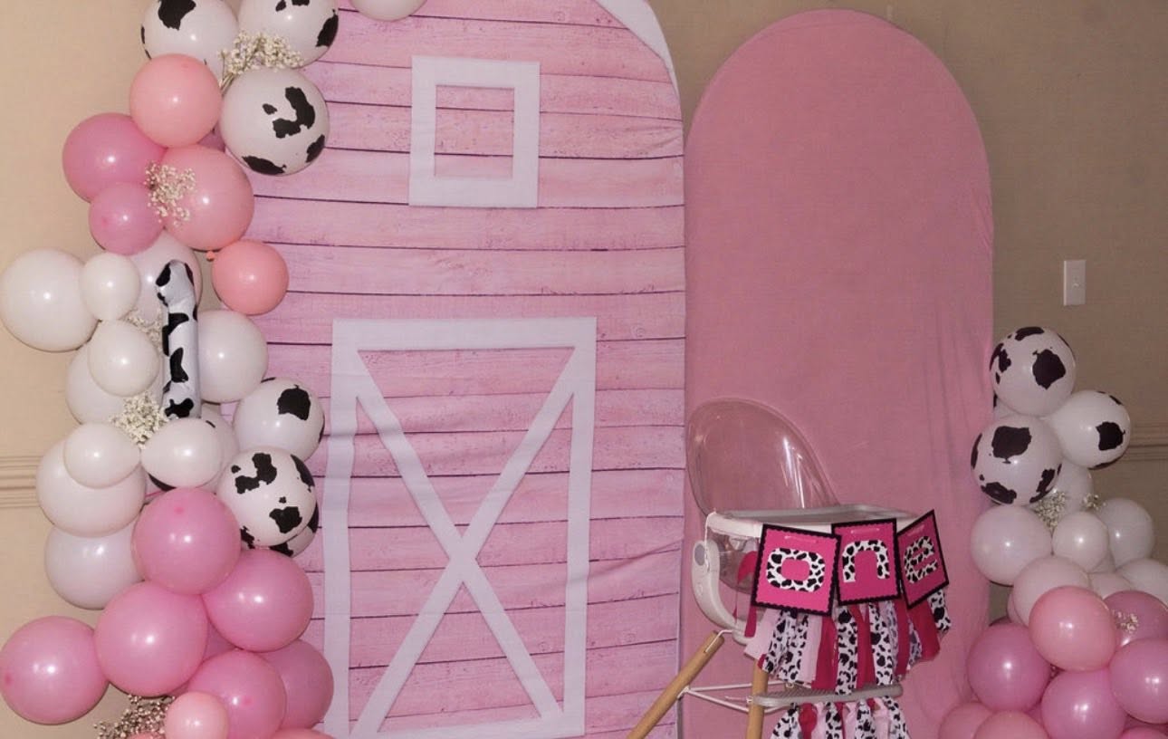Pink farm-themed birthday backdrop with balloon garlands, cow print accents, and a high chair for a first birthday
