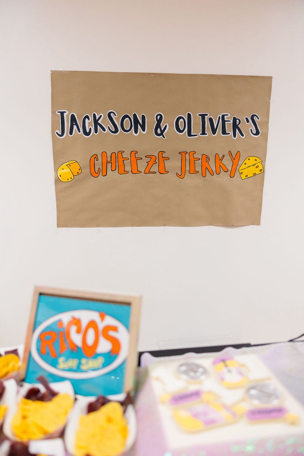 Homemade sign reading Jackson & Oliver's Cheeze Jerky above a snack station with Rico's Surf Shop sign.