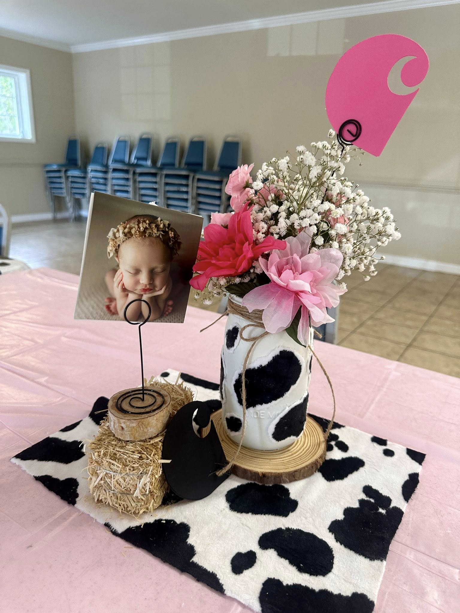 Cow print mason jar centerpiece with pink flowers, a baby photo placeholder, and miniature hay bale decor.