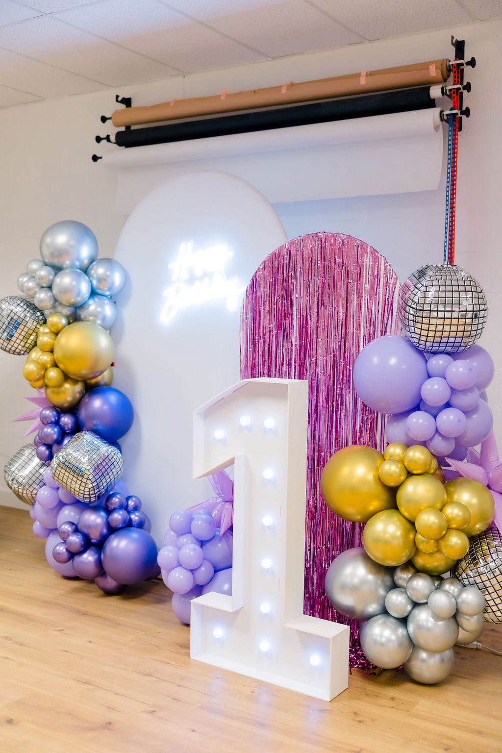 First birthday party backdrop with a light-up number one, balloons, disco balls, and a happy birthday neon sign.