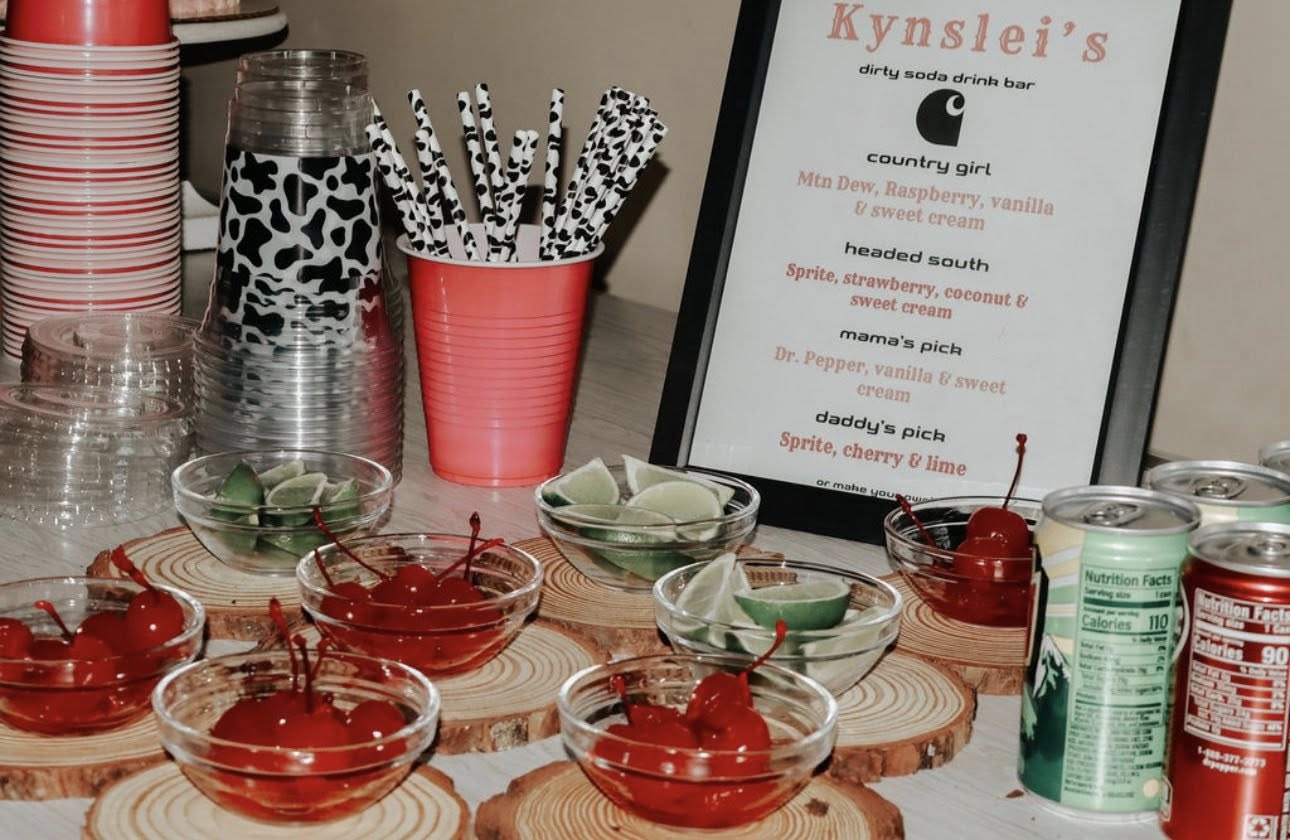 Table with a Kynslei's dirty soda bar sign, featuring bowls of maraschino cherries and lime wedges with cow-print straws.
