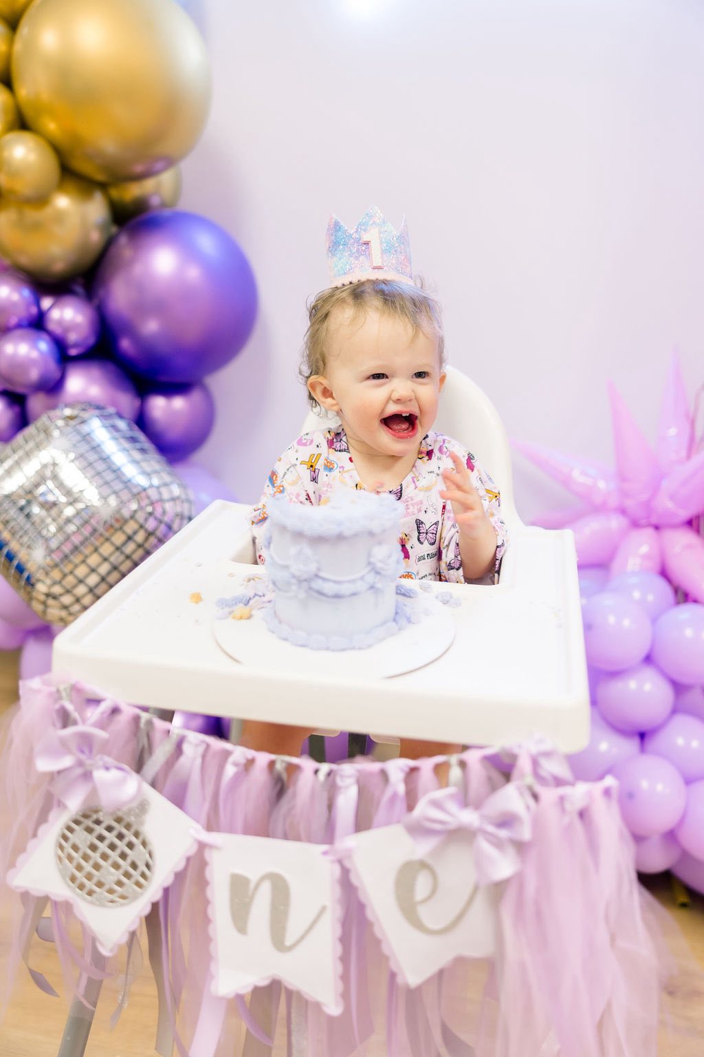 A laughing one-year-old child wearing a sparkly crown at a disco-themed first birthday party with purple balloons.