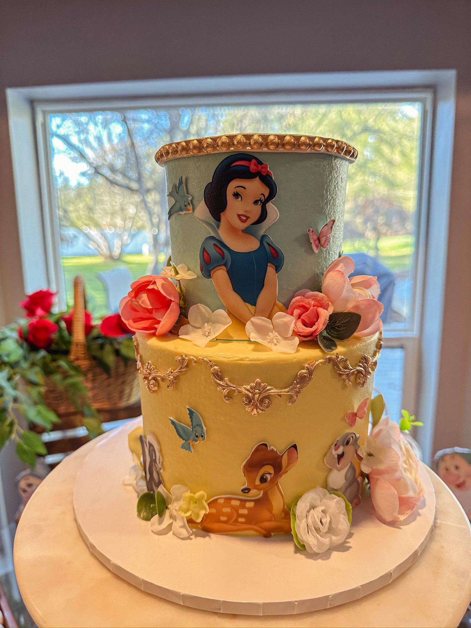 Two-tiered yellow cake featuring Snow White on top and forest animals including Bambi and Thumper on the base with floral decorations.