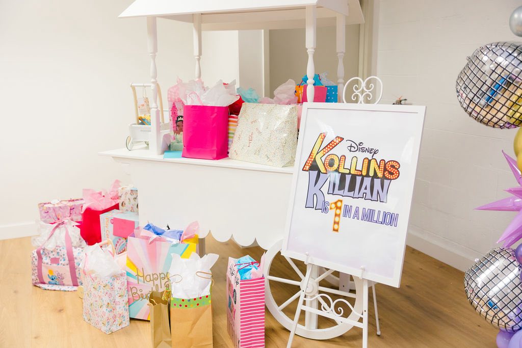 A white wooden party cart overflowing with colorful gift bags and a birthday sign reading Disney Kollins is 1 in a million.