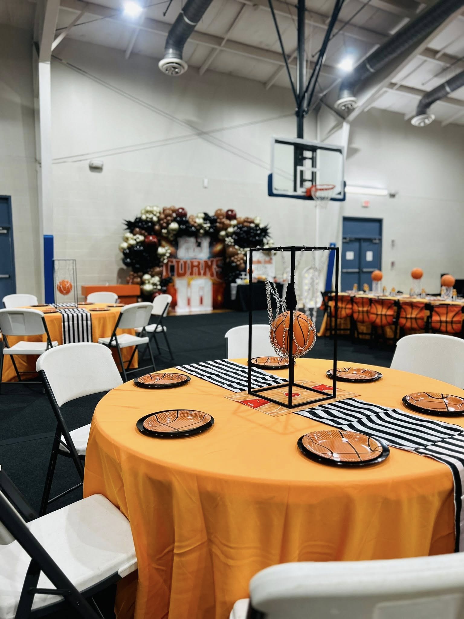 Basketball-themed party table settings with orange tablecloths and matching basketball plates in a gymnasium.