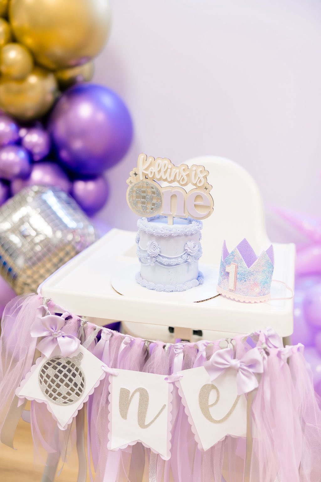 First birthday high chair setup with a purple cake, matching number one crown, and disco ball themed banner.