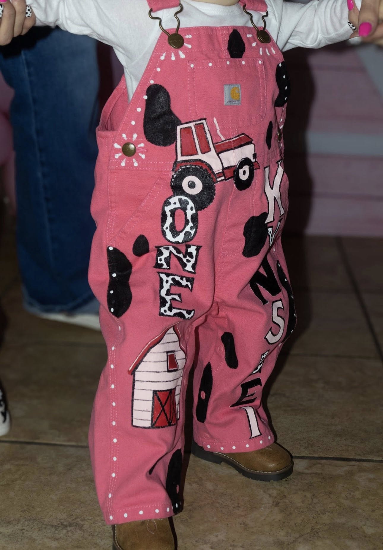 Pink Carhartt toddler overalls with custom farm-themed paint featuring a tractor, barn, and cow print patterns.