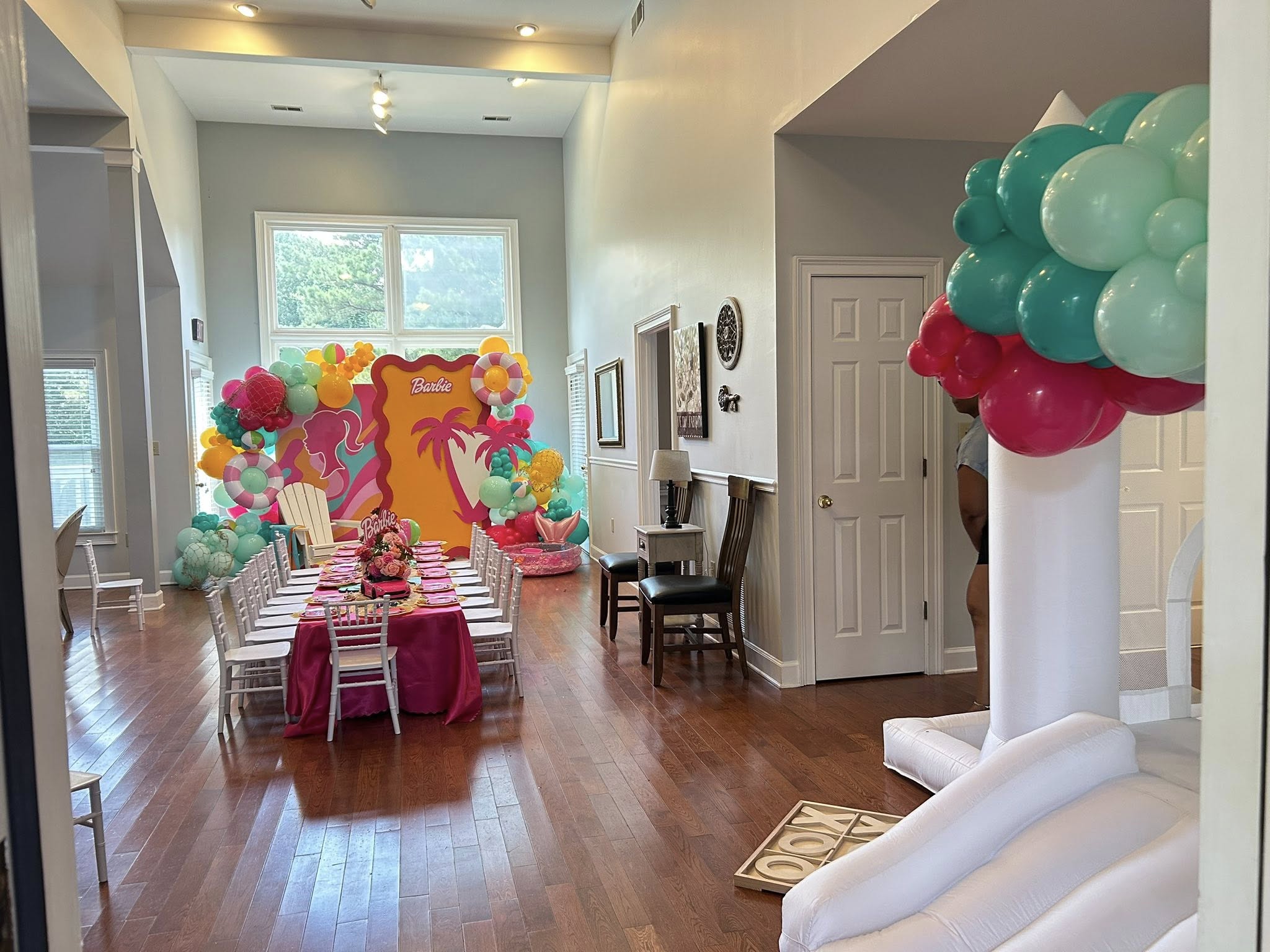 Living room decorated for a Barbie-themed party with a pink long table, chairs, and colorful balloon arch backdrop.