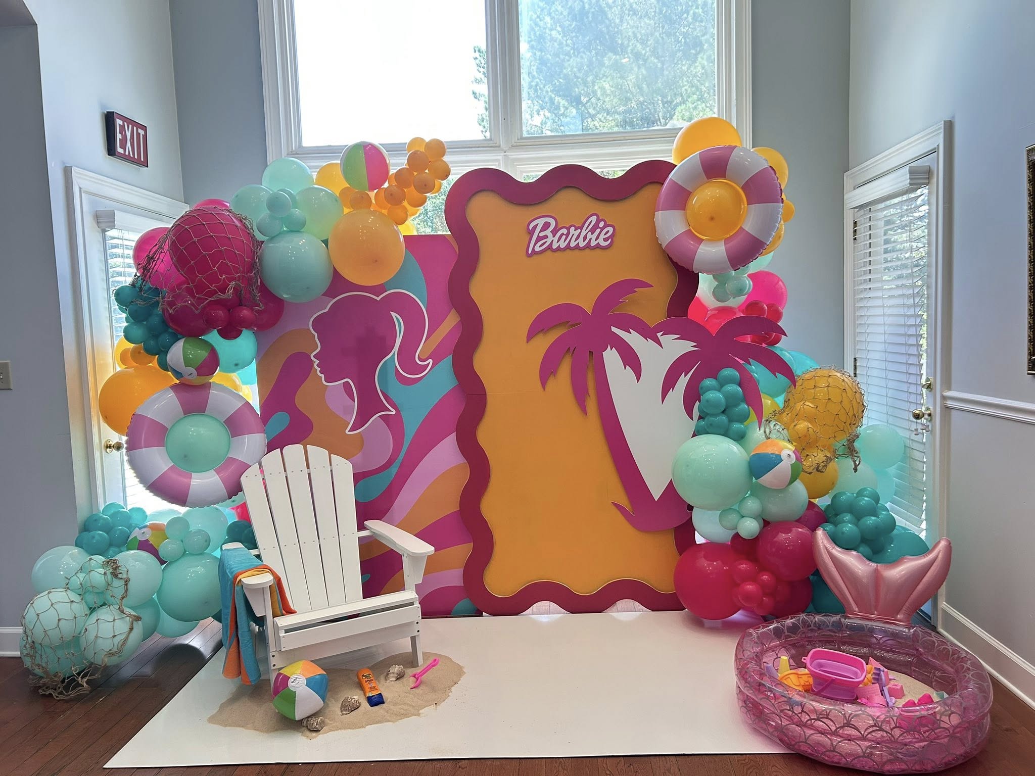 Barbie themed beach party photo backdrop featuring a white beach chair, balloons, and palm tree decorations.