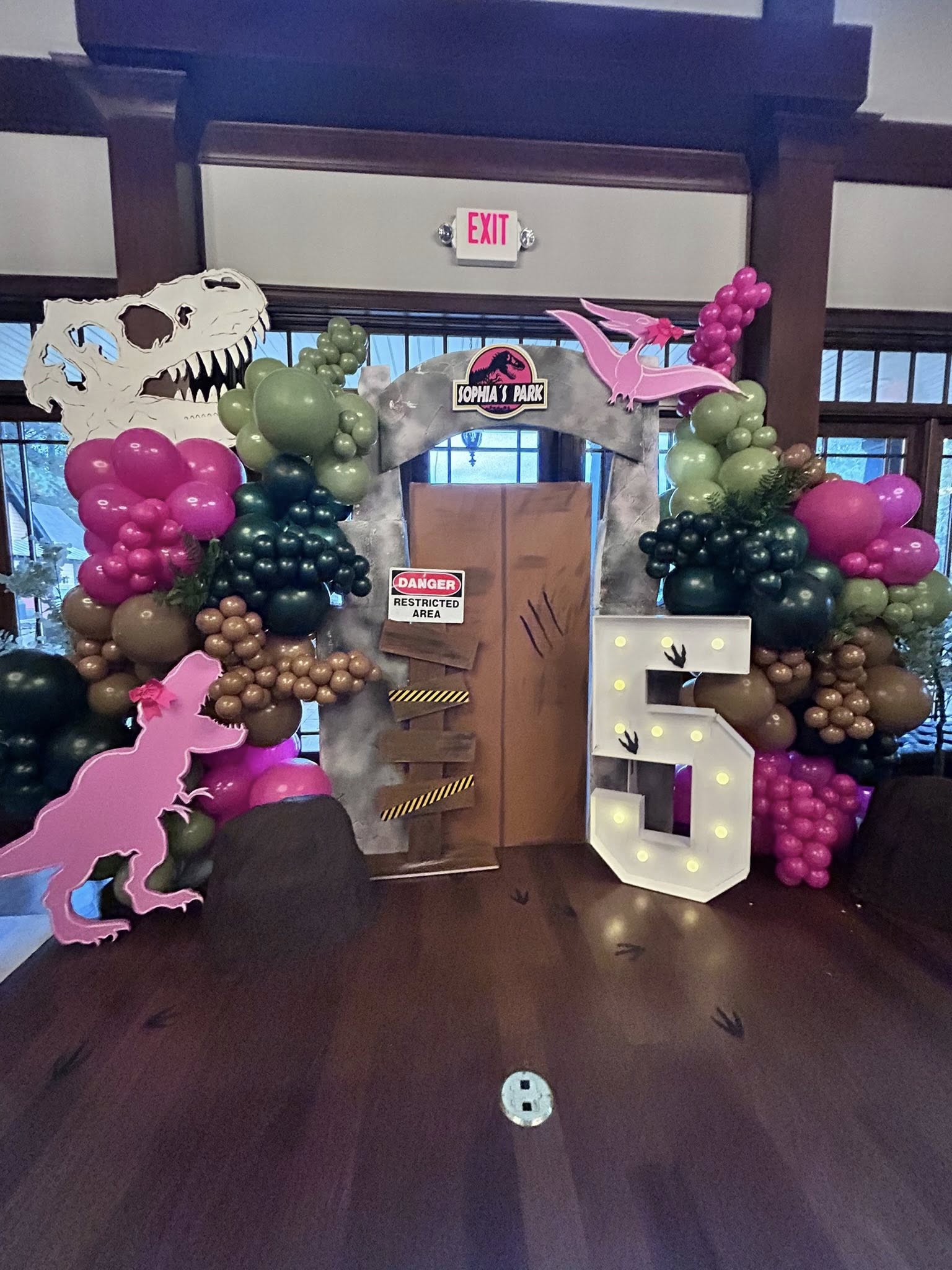 Sophia's Park dinosaur themed party backdrop with balloon arch, large number five light, and danger signage.