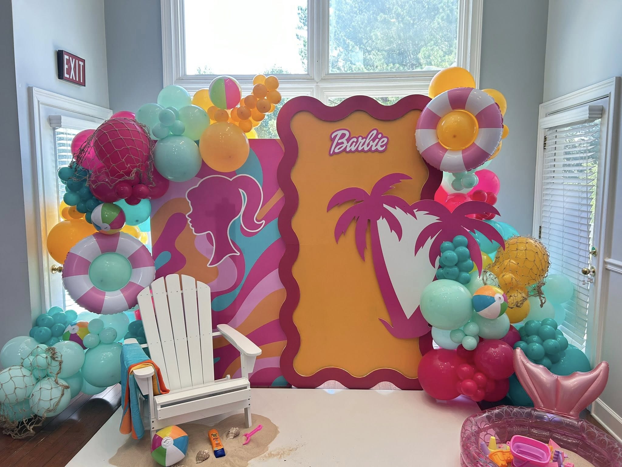 Barbie themed beach party photo backdrop with balloons, palm trees, and an Adirondack chair.
