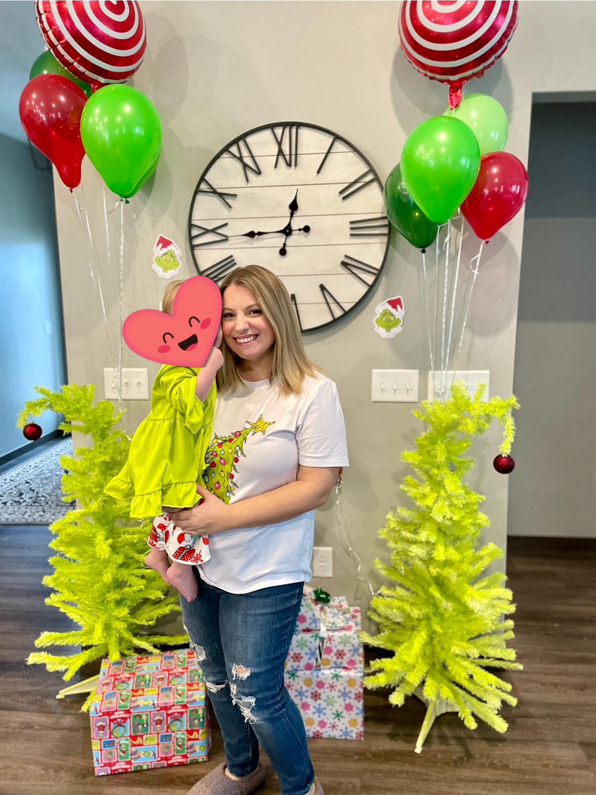 Woman wearing a Grinch-themed t-shirt posing with a toddler in front of Christmas decorations and balloons