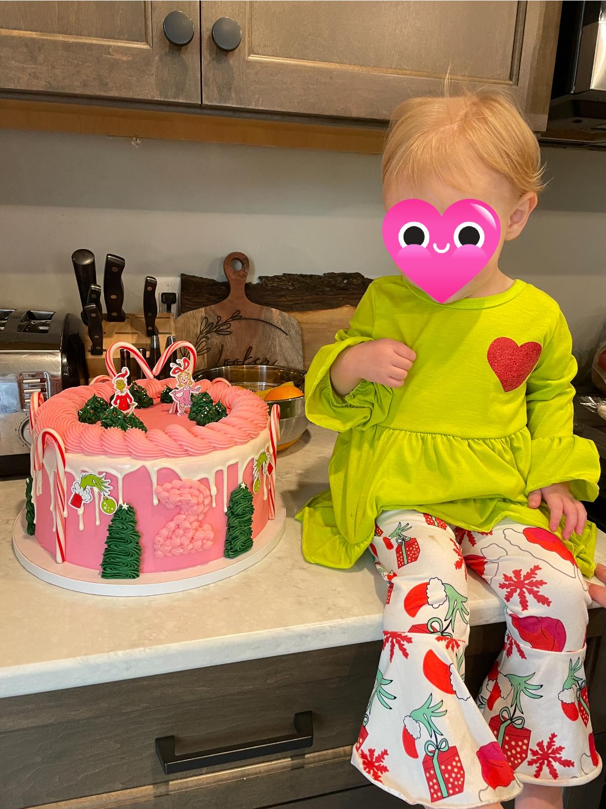 Toddler wearing a bright green top and Grinch-themed bell-bottom pants sitting next to a custom pink second birthday cake