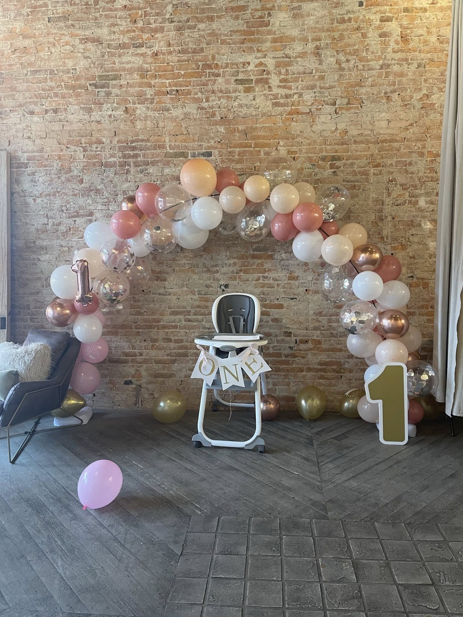 High chair for first birthday party decorated with a balloon arch and a One banner in front of a brick wall