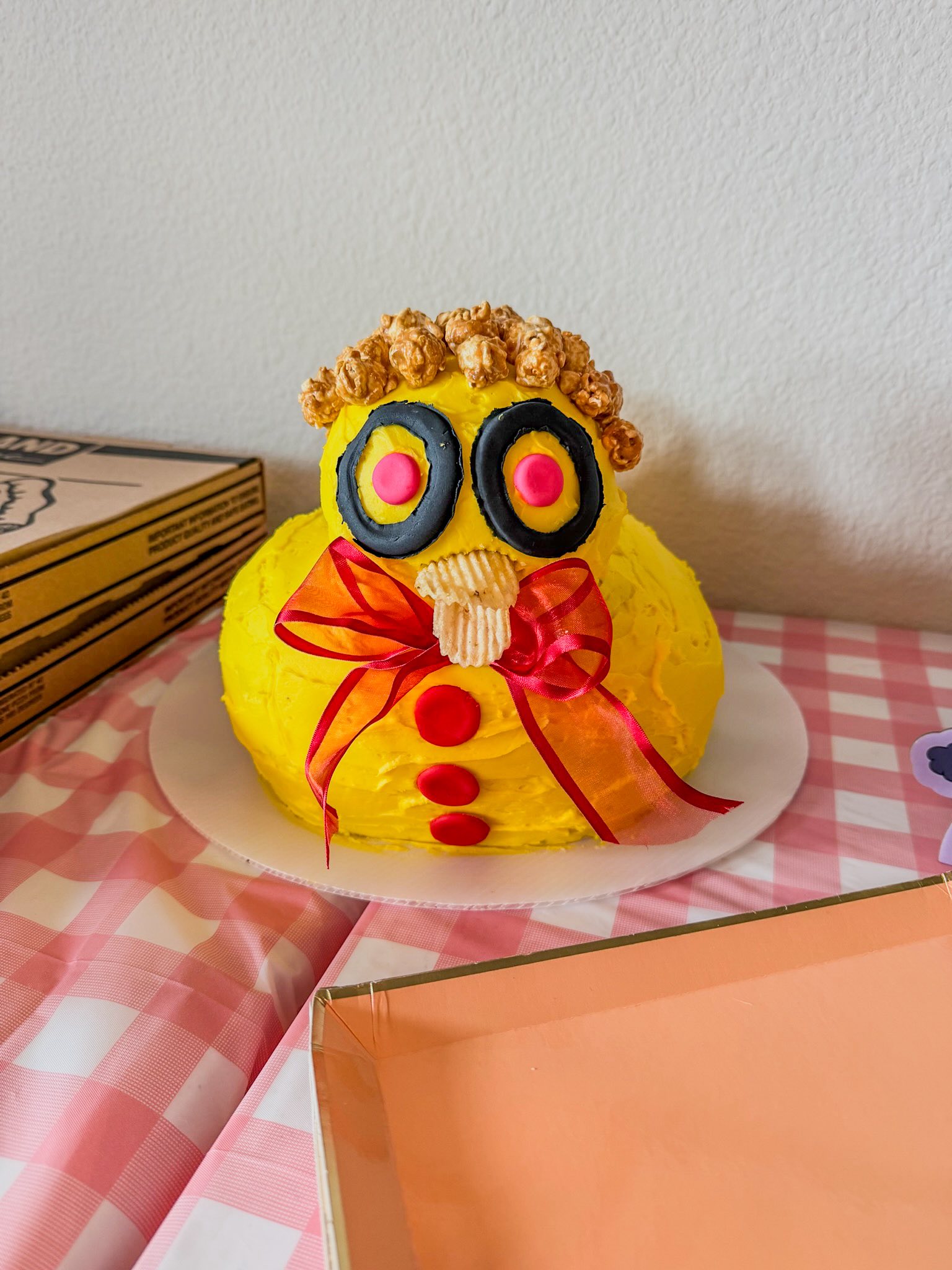 Yellow frosted cake decorated to look like a chicken with a red ribbon bowtie on a pink checkered tablecloth.