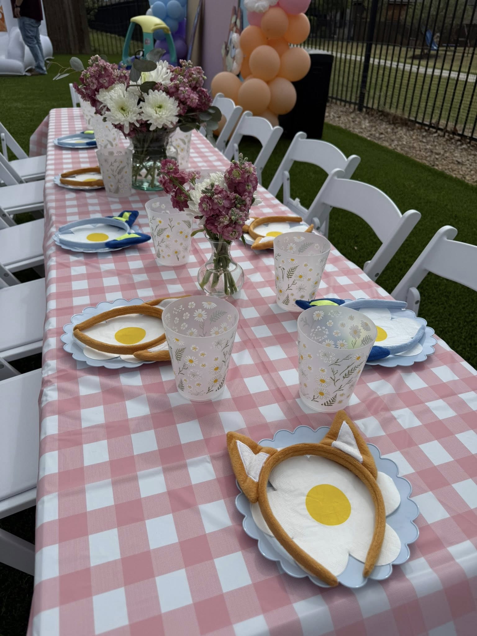 Outdoor birthday party table with pink gingham tablecloth, flower vases, and animal ear headbands on place settings.