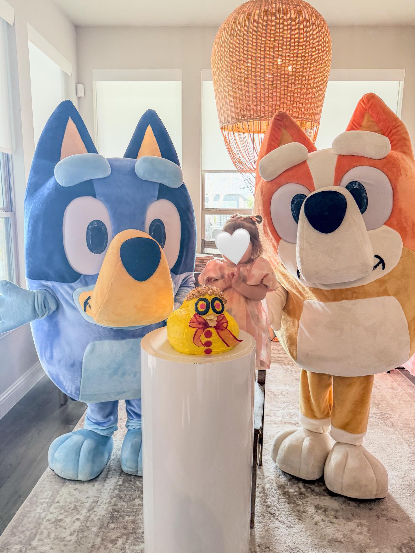 Bluey and Bingo character mascots standing with a small girl at a party with a duck-shaped cake.