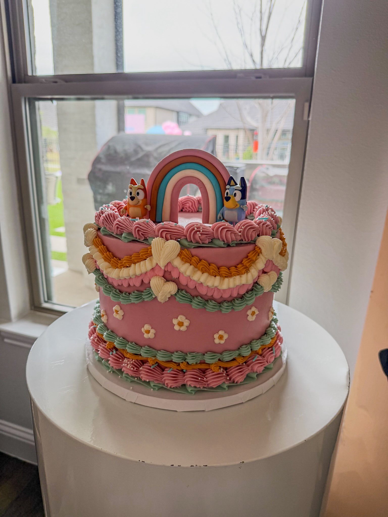 Pink tiered birthday cake decorated with Bluey and Bingo figurines, a rainbow topper, and piped buttercream details.