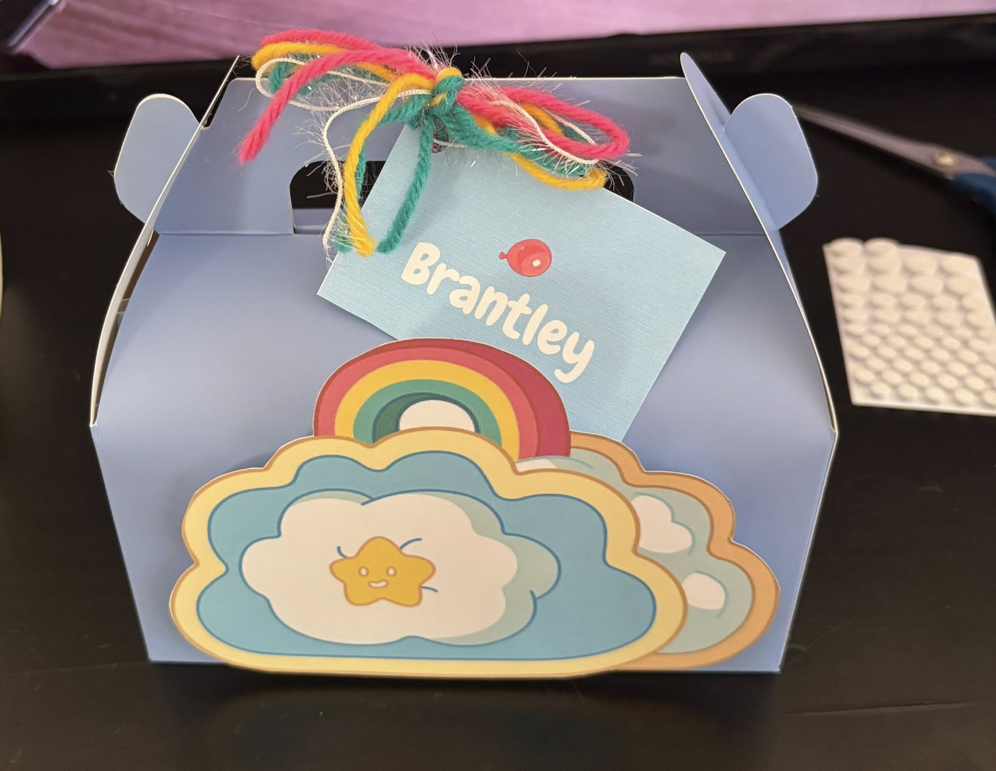 Light blue gift box decorated with a rainbow, cloud, and happy star topper, featuring a tag personalized with Brantley
